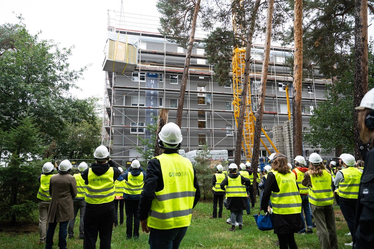 Quartier Jaminstraße Mehrere Personen mit Warnweste und Helm auf dem Kop auf einer Baustelle blicken zum schwebenden Bauelemente.