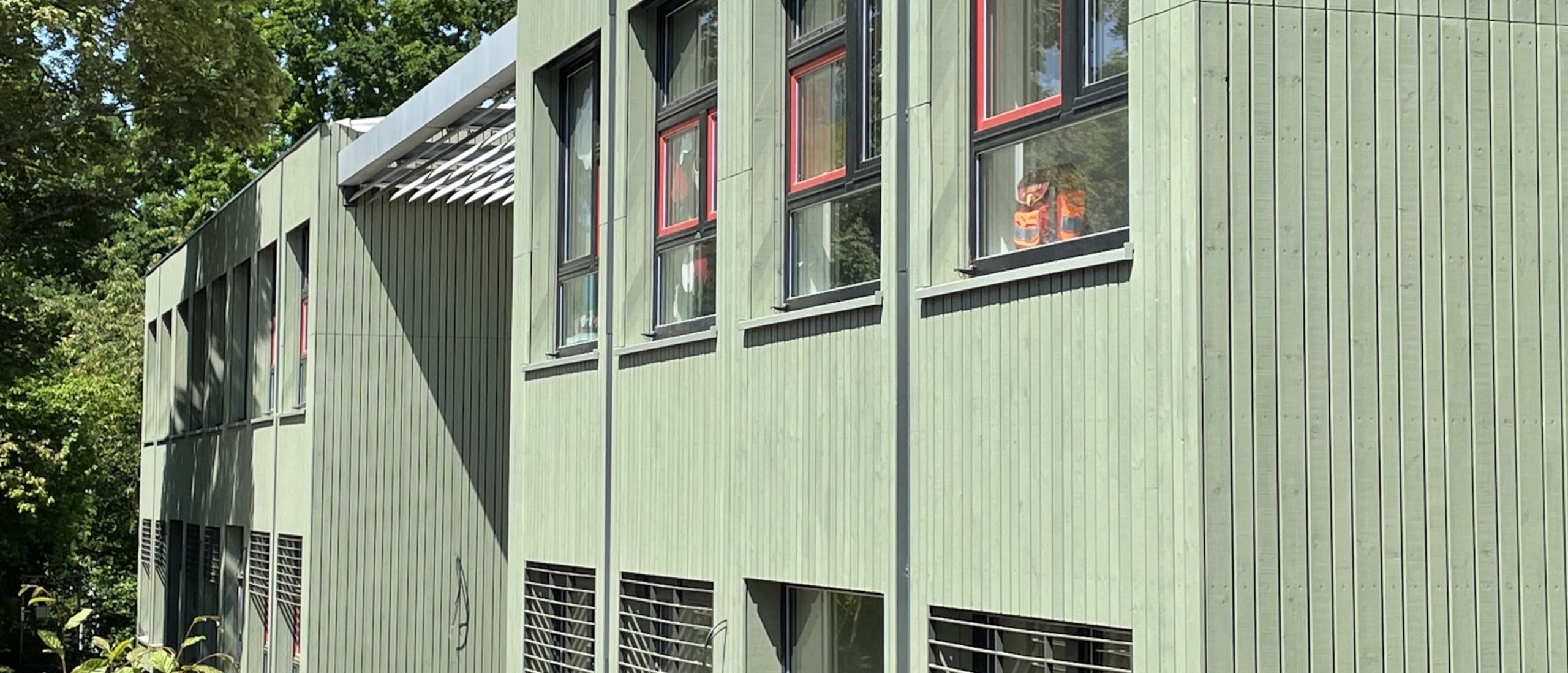 Kita-Gebäude zweigeschossig mit hellgrüner Holzfassade, blauer Himmel und Laubbäume