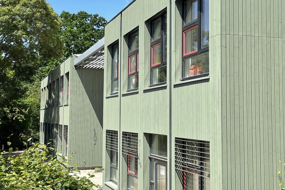 Verschieben Sie den Pfeil für den Vorher/Nachher-Vergleich Kita-Gebäude zweigeschossig mit hellgrüner Holzfassade, blauer Himmel und Laubbäume