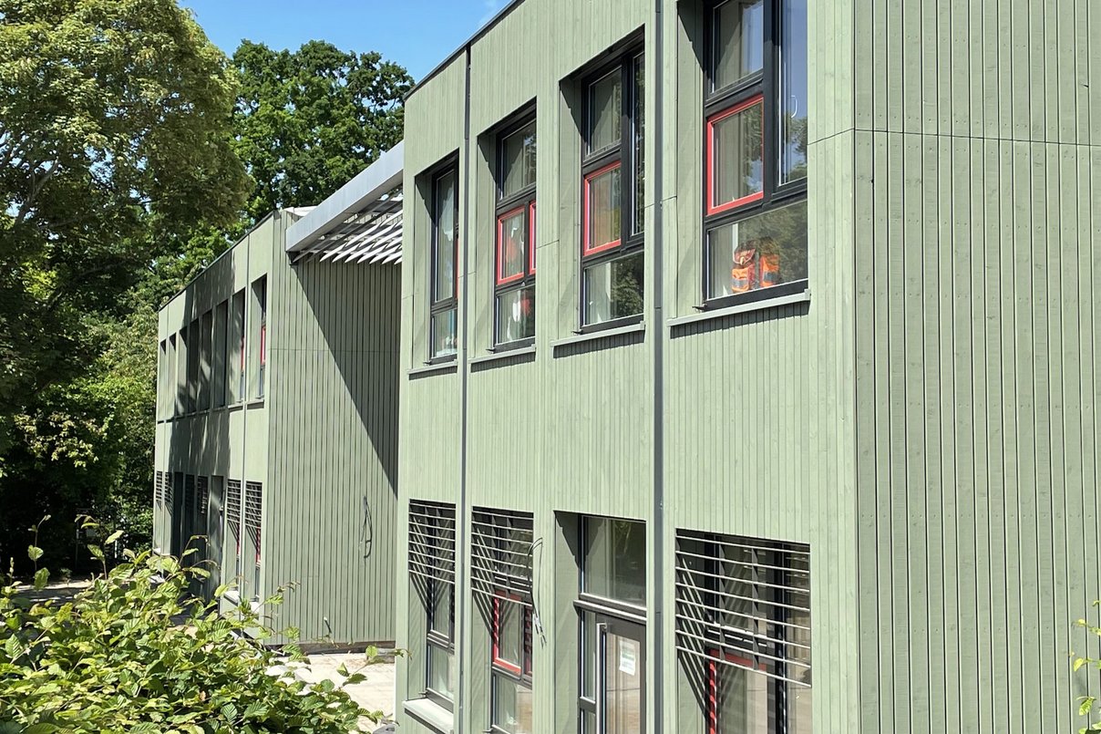 Kita-Gebäude zweigeschossig mit hellgrüner Holzfassade, blauer Himmel und Laubbäume