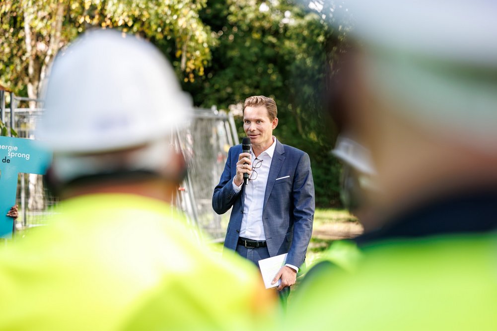 Bild zeigt Mann mit Mikro und im Vordergrund unscharf einen Mann mit Bauhelm und Weste
