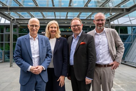 Vier Personen stehen nebeneinander für ein Gruppenbild, im Hintergrund Eingangsbereich eines Gebäudes