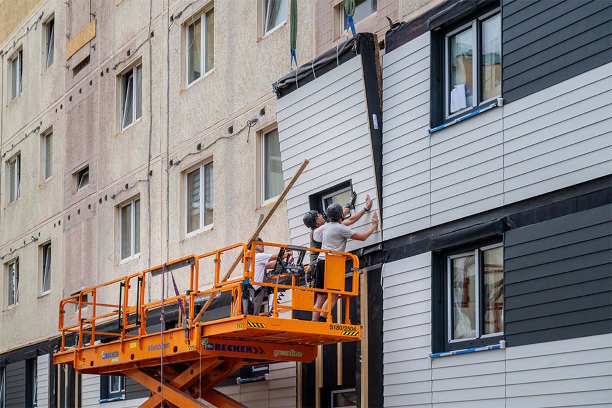 Nach der seriellen Sanierung erreicht der Plattenbau in der Zerbster Straße KfW-Effizienzhausstandard 40EE und ist damit energetisch auf Neubau-Niveau. Drei Bauarbeiter stehen auf einer Hebebühne und befestigen ein vorgefertigtes Hausfassadenelement.