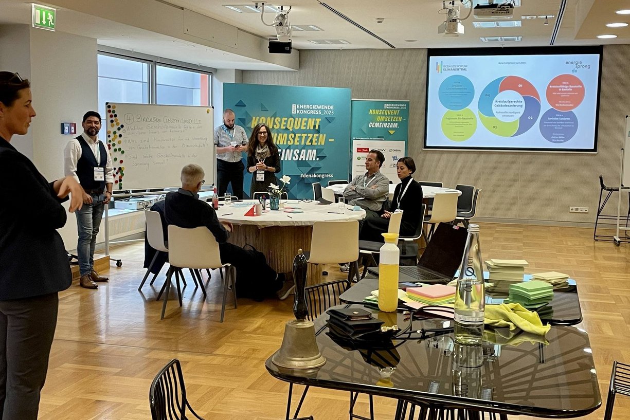 An vier Tischen wurden vier Themen rund um die kreislaufgerechte Bestandssanierung diskutiert und im Austausch vielversprechende Ideen entwickelt. Workshopraum mit Tischen und Gruppe an Arbeitenden in Diskussion