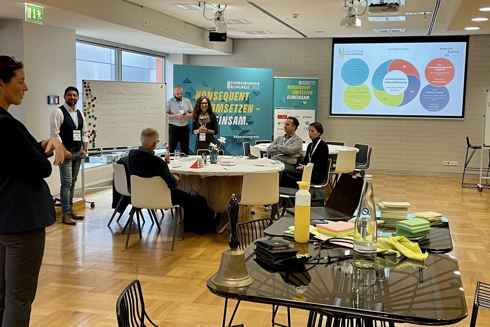 An vier Tischen wurden vier Themen rund um die kreislaufgerechte Bestandssanierung diskutiert und im Austausch vielversprechende Ideen entwickelt. Workshopraum mit Tischen und Gruppe an Arbeitenden in Diskussion