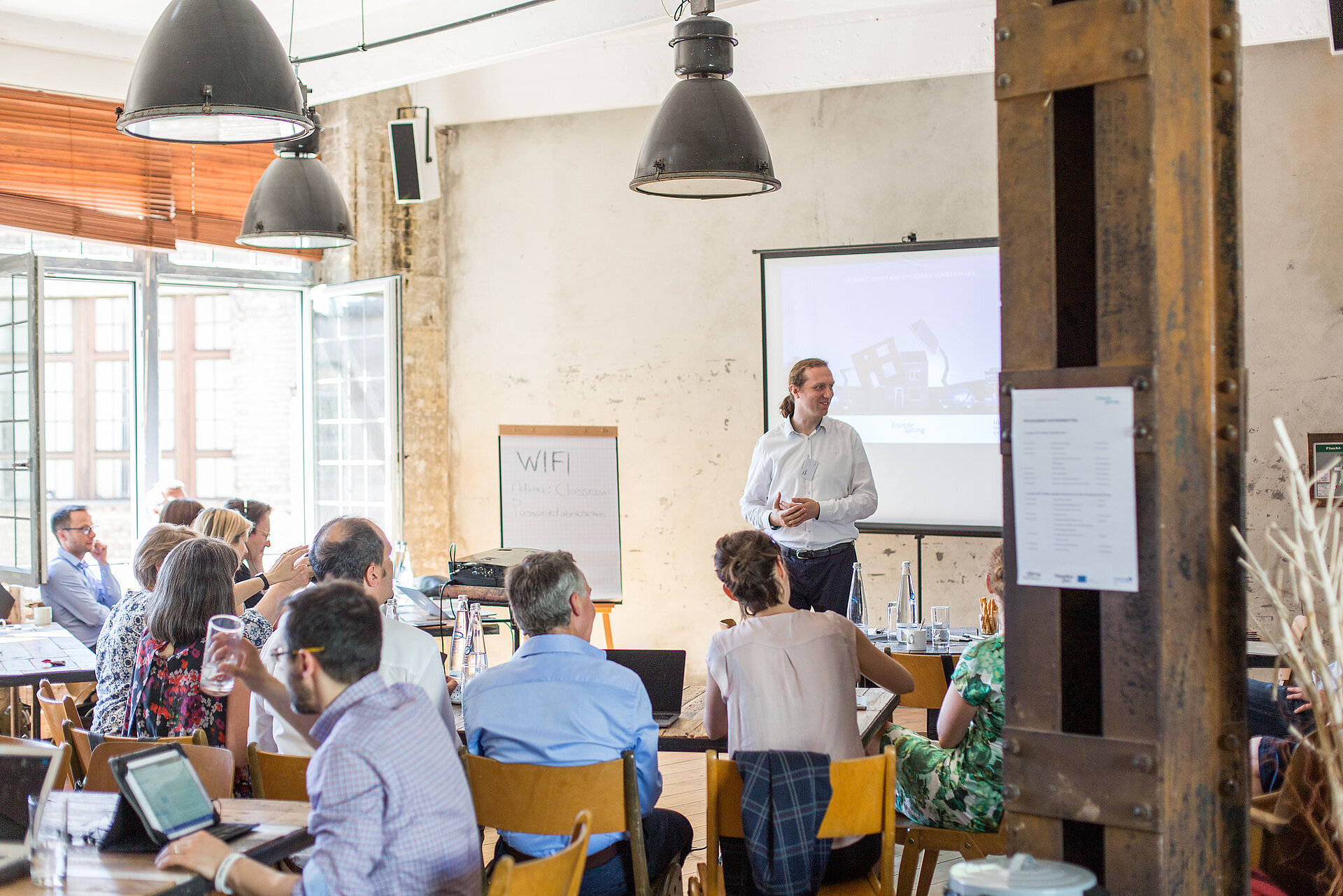 Personen sitzen in einem Raum, vorne steht eine Person, im Hintergrund sind eine Projektionsleinwand und ein Flipchart.