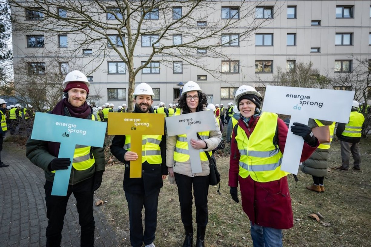 Vier Personen mit Bauhelmen auf dem Kopf stehen nebeneinander. In der Hand halten sie jeweils ein Schild mit der Aufschrift "energiesprong.de".