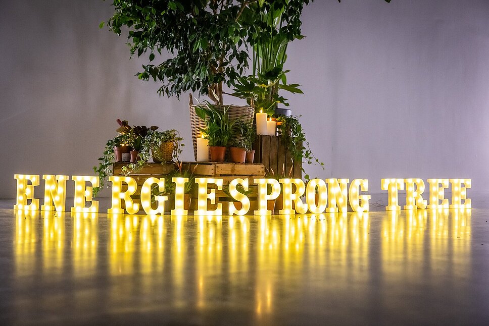 Auf dem Boden stehen nebeneinander gelb leuchtende Buchstaben und ergeben die Wort: ENERGIESPRONG TREE. Dahinter stehen Topfpflanzen.