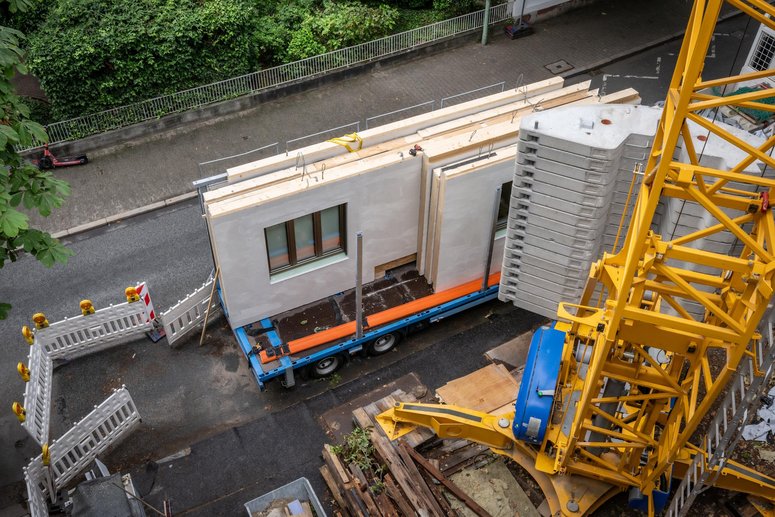 Blick von oben auf einen Baustellenbereich mit Kran und Anhänger, der vorgefertigte Hausfassadenelemente geladen hat.