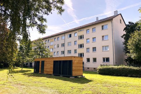 Bild zeigt Container eines Energiemoduls auf grüner Wiese vor einem Mehrfamilienhaus.