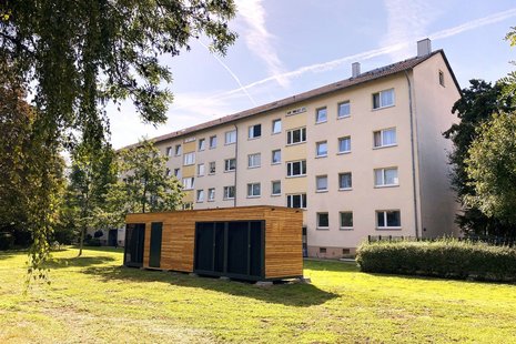 Bild zeigt Container eines Energiemoduls auf grüner Wiese vor einem Mehrfamilienhaus.