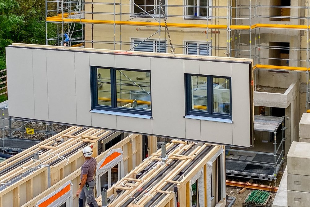 Serielle Sanieren setzt auf digitale Planung, modulare Vorfertigung und standardisierte Prozesse. Bestandsgebäude lassen sich so mit weniger Fachkräften schneller auf den klimaneutralen NetZero-Standard bringen. Schwebendes Fassadenelement mit Fenstern; Bauarbeiter, der auf weiteren steht