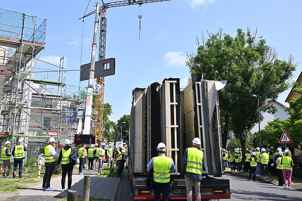 Mehrere Personen vor einer Baustelle, auf der Straße ein LKW mit vorgefertigten Haus-Fassadenelementen