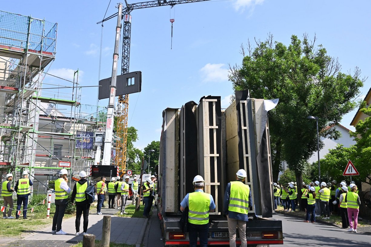 Mehrere Personen vor einer Baustelle, auf der Straße ein LKW mit vorgefertigten Haus-Fassadenelementen