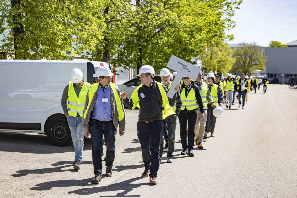 Bild zeigt: Menschenschlange mit Bauwesten und Bauhelmen, vorne wird ein Helm hochgehalten.