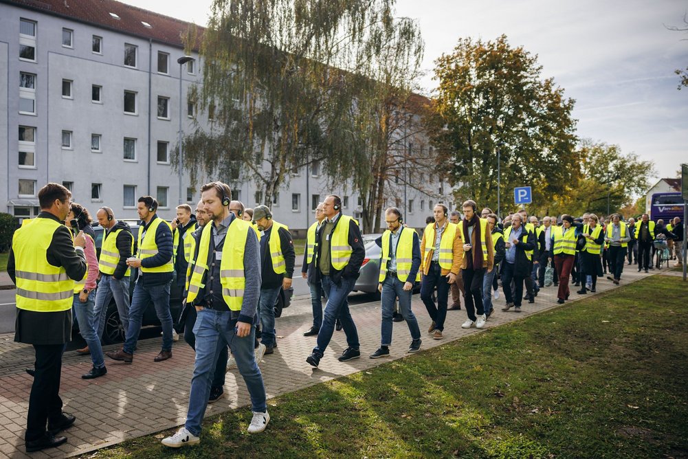 Mehrere Personen in gelben Sicherheitswesten laufen hintereinander auf einem FUßweg, im Hintergrund ist ein Mehrfamilienhaus.
