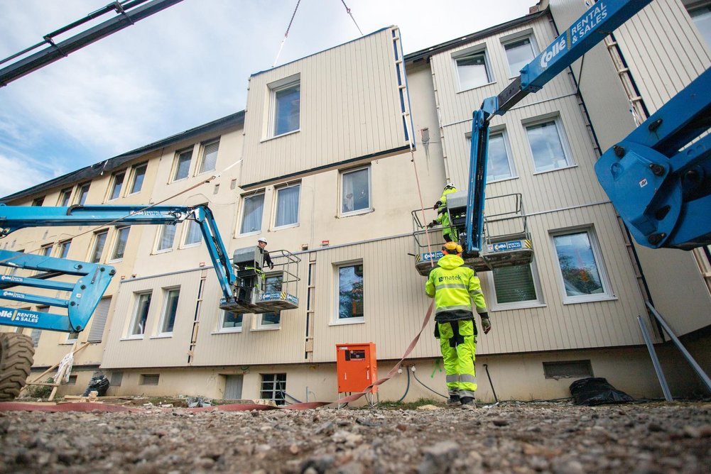 Renowate und Matek – ein eingespieltes Team. Das LEG Joint Venture und der estländische Holzbauspezialist haben bereits zwei Quartiere in Mönchengladbach seriell saniert. Arbeiter bringen per Kran vorgefertigte Fassadenteile an Gebäude an