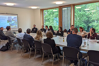 Personen sitzen an einem großen weißen Tisch in einem Raum mit großem Fenster.