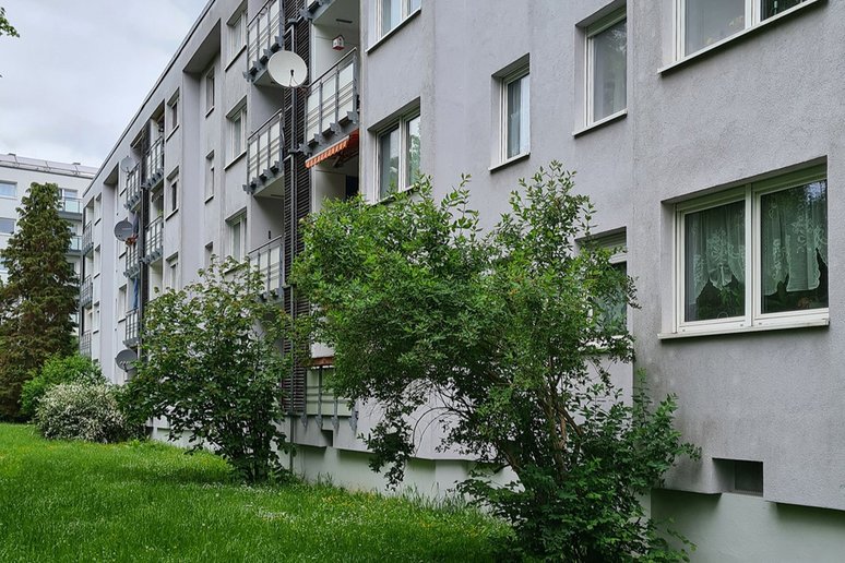 Gartenseite eines mehrgeschossigen Mehrfamilienhauses mit hellgrauer Fassade. Entlang des Gebäudes befinden sich mehrere Balkone mit Metallgeländern und Satellitenschüsseln. Davor wachsen dicht bepflanzte Sträucher und Bäume auf einer grünen Rasenfläche.