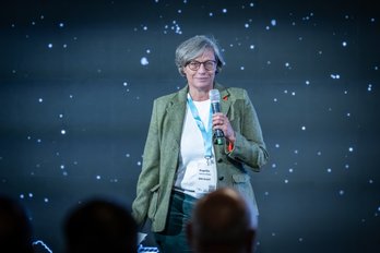 Person steht mit Mikrofon in der Hand vor Publikum.