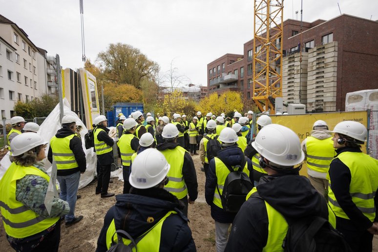 Personen in gelben Sicherheitswesten und mit weißen Bauhelmen auf dem Kopf stehen auf einer Baustelle, im Hintergrund Wohngebäude.