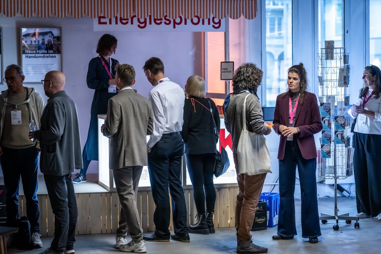 Personen stehen an einem Kiosk auf einer Veranstaltung und sind teils in Gesprächen.