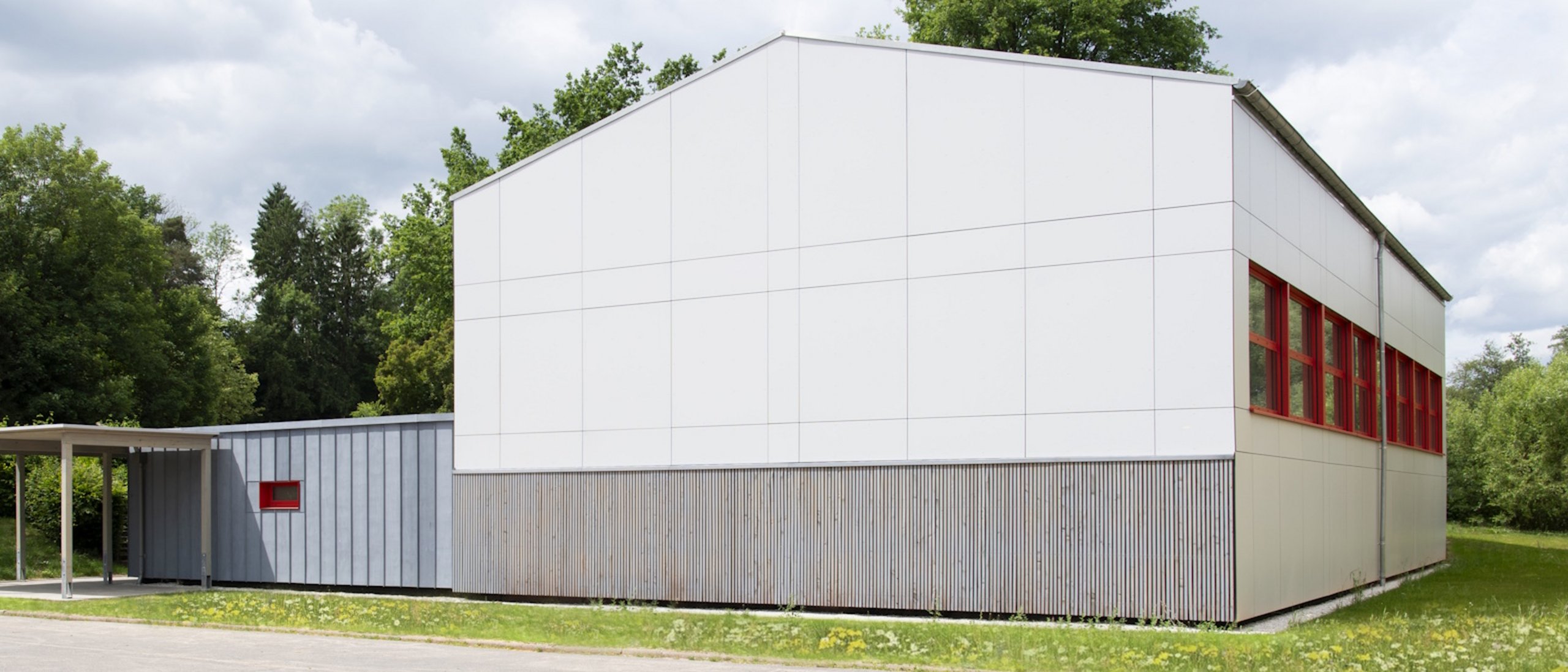 Außenansicht neu sanierte Turnhalle umgeben von Rasenflächen und Laubbäumen, davor eine große betonierte Fläche