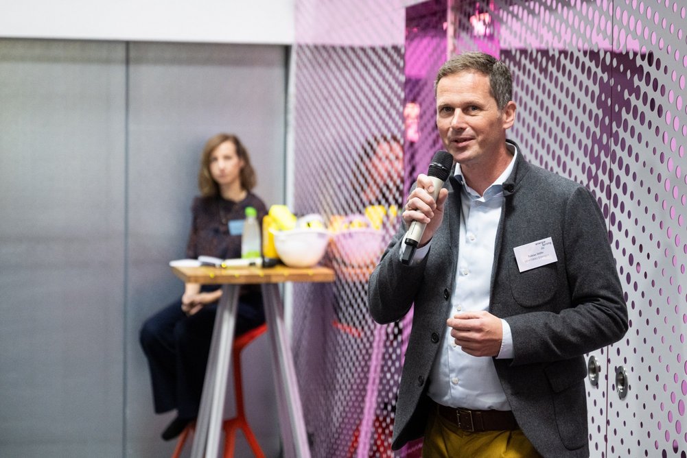 Tobias Stöhr, Geschäftsführer der GEWOBAU Erlangen Person im Anzug mit Mikrofon in der Hand, im Hintergrund sitzt eine Person an einem Stehtisch.