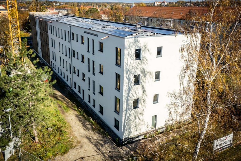 Blick von oben auf ein viergeschossiges Wohngebäude mit heller Putzfassade und Flachdach mit Photovoltaikanlage. Drumherum stehen herbstliche Laubbäume.