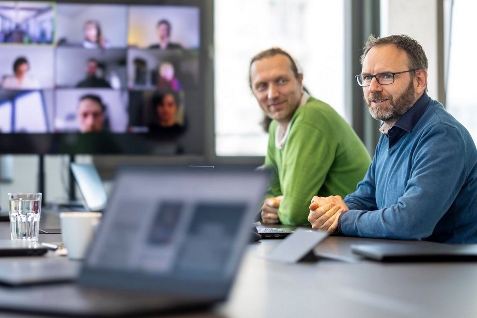 Zwei Personen sitzen nebeneinander am Tisch, im Hintergrund ein Bildschirm, im Vordergrund liegt ein Notebook auf dem Tisch