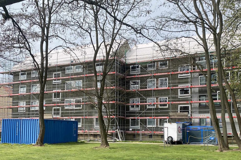 Im vordergrund Rasen und Laubbäume, im Hintergrund ein Mehrfamilienhaus (viergeschossig) mit Baugerüst