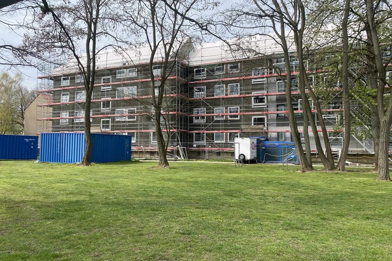 Im vordergrund Rasen und Laubbäume, im Hintergrund ein Mehrfamilienhaus (viergeschossig) mit Baugerüst