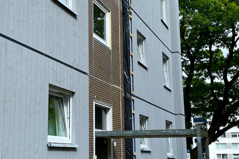 Mehrstöckiges Wohngebäude mit Gerüst und schwarzer Abdeckung an der Fassade, umgeben von einem gepflasterten Weg und Grünflächen unter klarem Himmel.