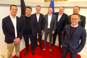 Sieben Personen in Business-Outfits stehen nebeneinander auf einem roten Teppich für ein Gruppenbild.