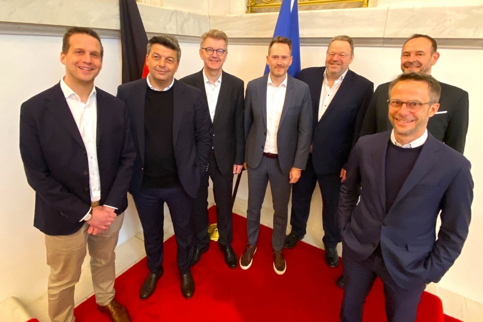 Sieben Personen in Business-Outfits stehen nebeneinander auf einem roten Teppich für ein Gruppenbild.