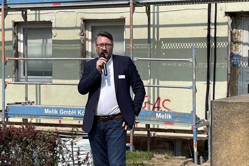 Guido Sinn, Technischer Vorstand der BWB Person im Anzug mit Mikrofon in der Hand, im Hintergrund eine Hausfassade mit Baugerüst