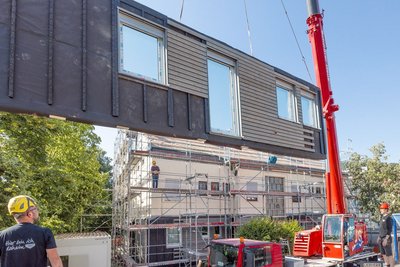Schwebendes Fassadenelement mit Fenstern an Kranfahrzeug, drei Bauarbeiter schauen stehen und schauen
