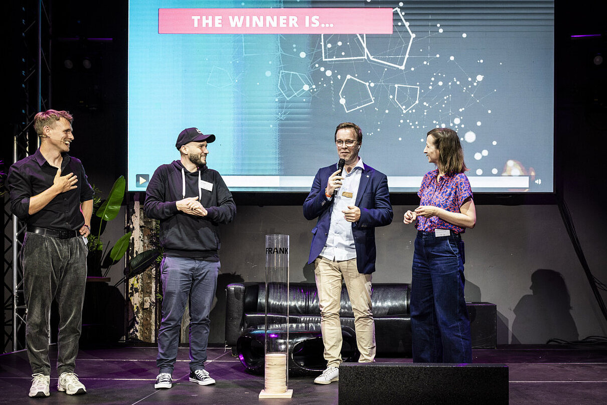 Vier Personen auf einer Bühne. Im Hintergrund eine Präsentation mit dem Text: The winner is ...