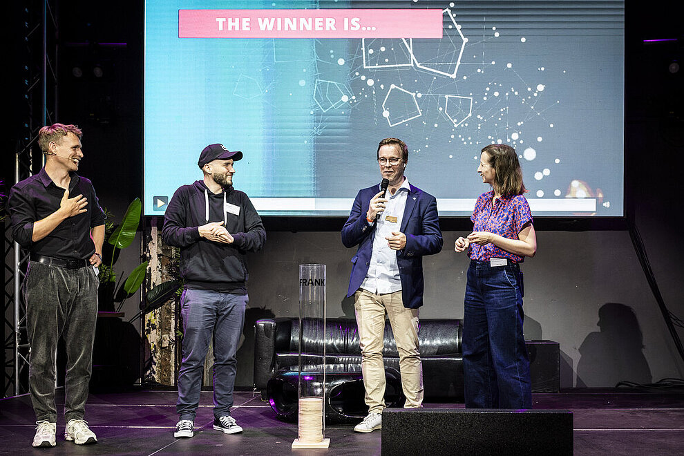 Vier Personen auf einer Bühne. Im Hintergrund eine Präsentation mit dem Text: The winner is ...