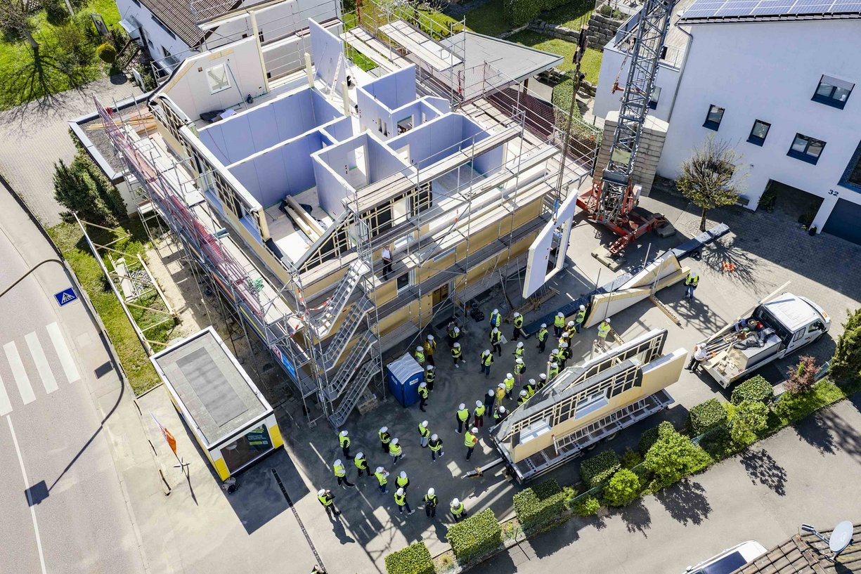 Bild zeigt: Baustelle mit Kran an dem Fassadenelemente fliegen, davor eine Menschentraube mit Bauhelmen und Bauwesten