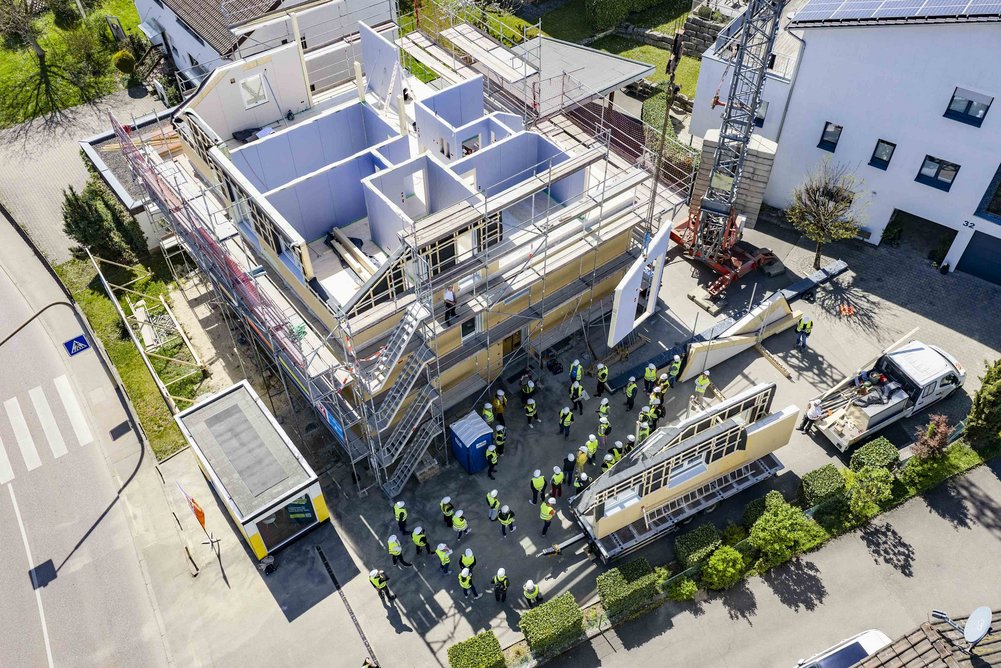 Bild zeigt: Baustelle mit Kran an dem Fassadenelemente fliegen, davor eine Menschentraube mit Bauhelmen und Bauwesten