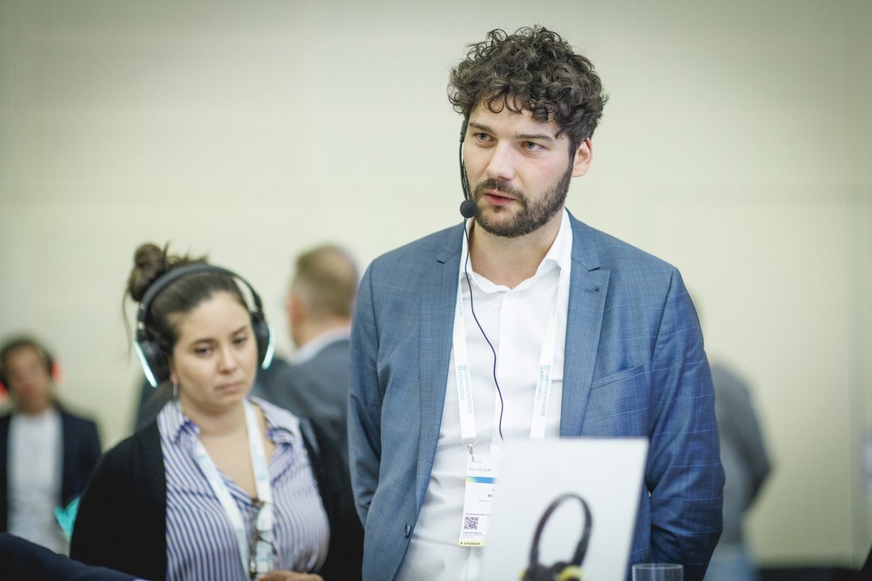 Marius Mersinger, Gründer und Geschäftsführer der Moducon Person mit Headset im Raum mit weiteren Personen, dahinter eine Person mit Kopfhörer