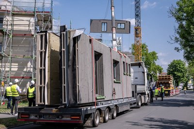 Transport mit vorgefertigten Hausfassenelementen steht auf einer Straße, im Hintergrund Mehrfamlienhäuser und ein Kran, an dem ein Fassadenelement hängt. Außerdem Personen mit gelben Sicherheitswesten und weißen Helmen sowie Laubbäume.