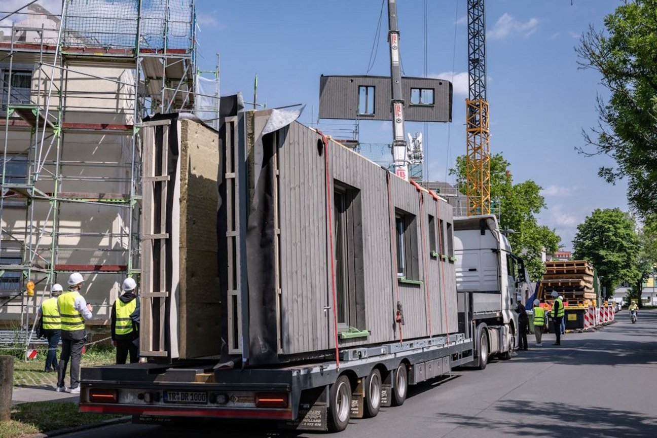 Transport mit vorgefertigten Hausfassenelementen steht auf einer Straße, im Hintergrund Mehrfamlienhäuser und ein Kran, an dem ein Fassadenelement hängt. Außerdem Personen mit gelben Sicherheitswesten und weißen Helmen sowie Laubbäume.