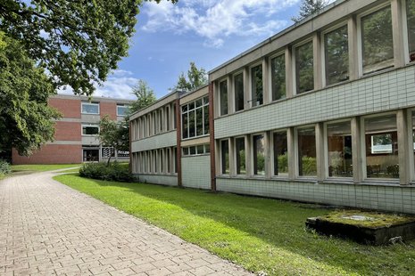 Gebäude mit großen Fenstern, zweigeschossig, unsaniert, davor Rasenfläche und ein Weg