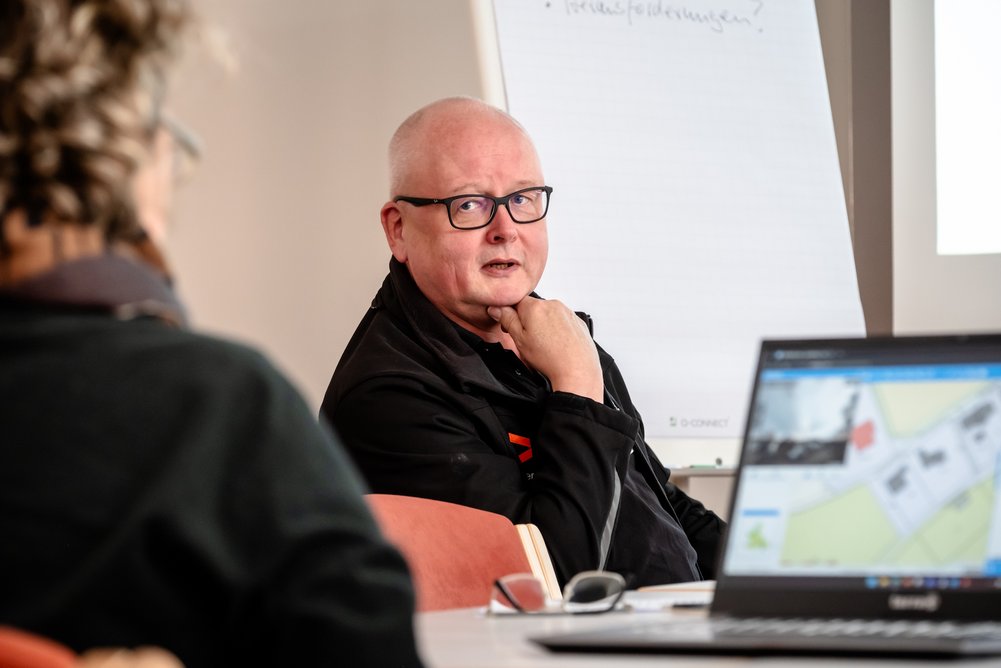 Person sitzt und schaut zu einer anderen Person. Im Hintergrund ein Flipchart, im Vordergrund ein Notebook.