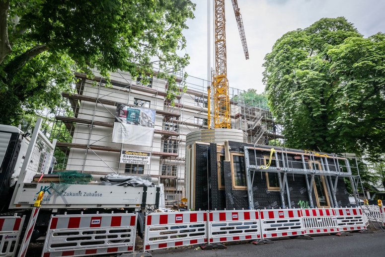 Bauabsperrung vor einem Wohngebäude mit Baugerüst, davor ein Kran und seitlich große Laubbäume
