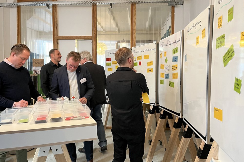 Mehr als 20 Teilnehmende aus den Bereichen Bau, Planung und Materialentwicklung arbeiteten interaktiv Kriterien für serielle Sanierungsstandards aus. Fünf Personen stehen in einem Workshop-Raum, Stellwände mit farbigen Post-its darauf