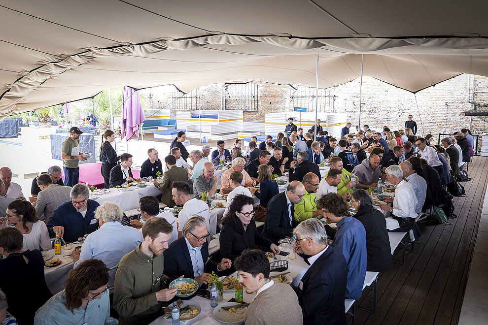 Viele Menschen sitzen an langen gedeckten Tischen und essen. Darüber ist ein Zeltdach gespannt.