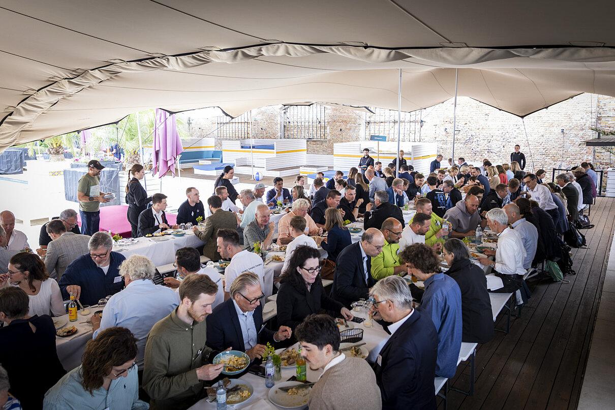 Viele Menschen sitzen an langen gedeckten Tischen und essen. Darüber ist ein Zeltdach gespannt.